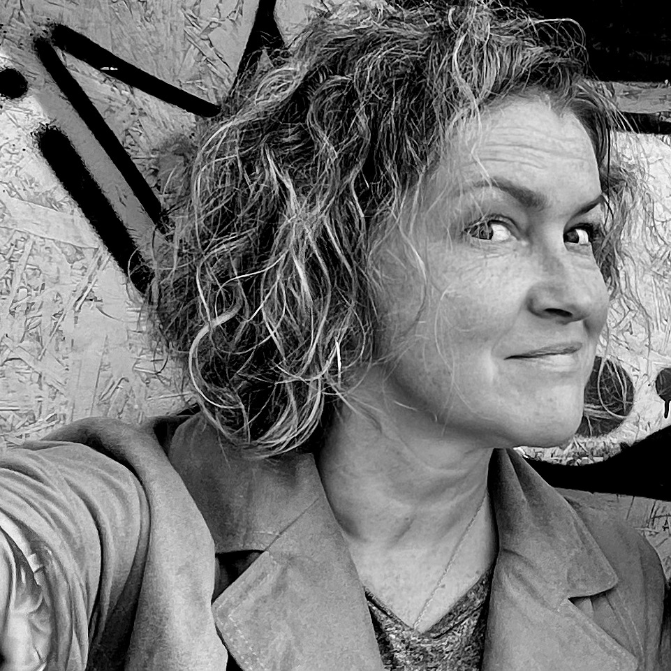 Cecily Salt portrait 2024 in black and white against a chipboard graffiti wall at Notting Hill Carnival. She has shoulder length curly hair with highlights, looking to camera